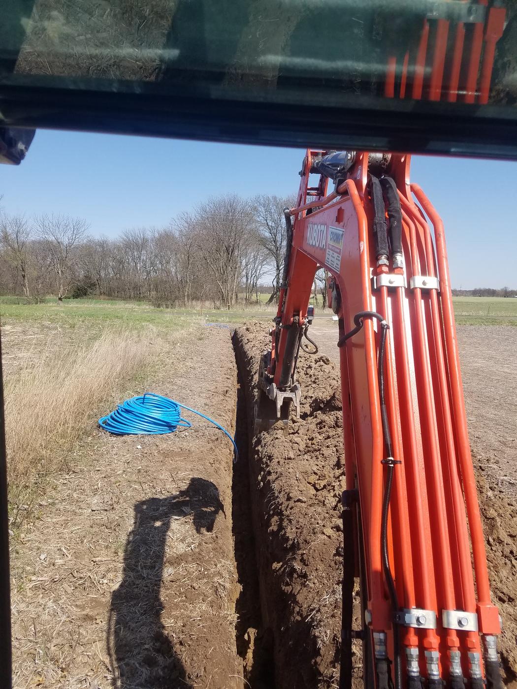 A coil of blue water service pipe beside a freshly cut trench, seen from inside the excavator cab