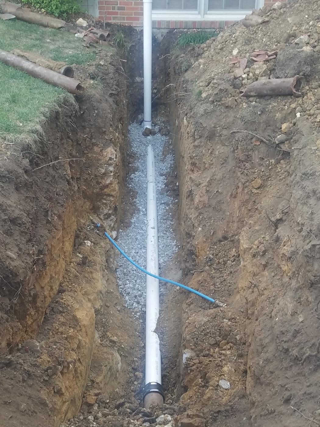 A sewer line replacement: new white PVC bedded on gravel in a deep trench beside a brick house, with the old corroded cast iron dug out on the lawn above
