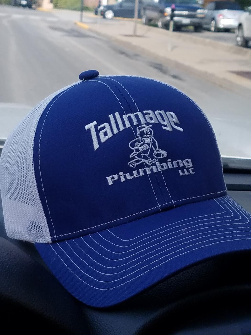 A Tallmage Plumbing cap on a dashboard, royal blue with the wordmark and mascot embroidered in white